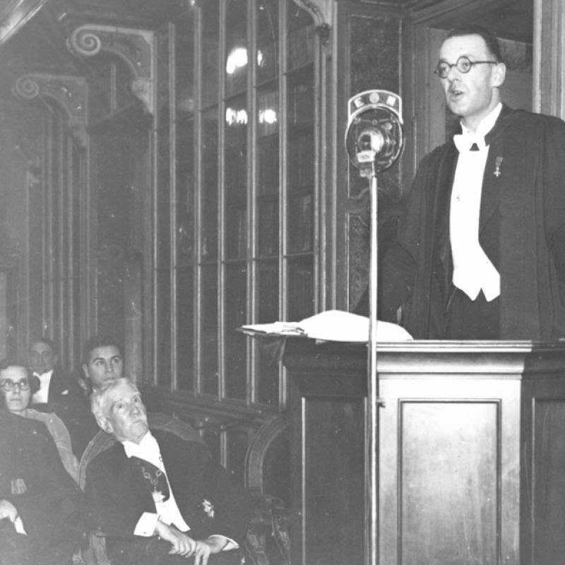 Professor West standing at a lectern addressing a smartly dressed audience in an ornately decorated room.