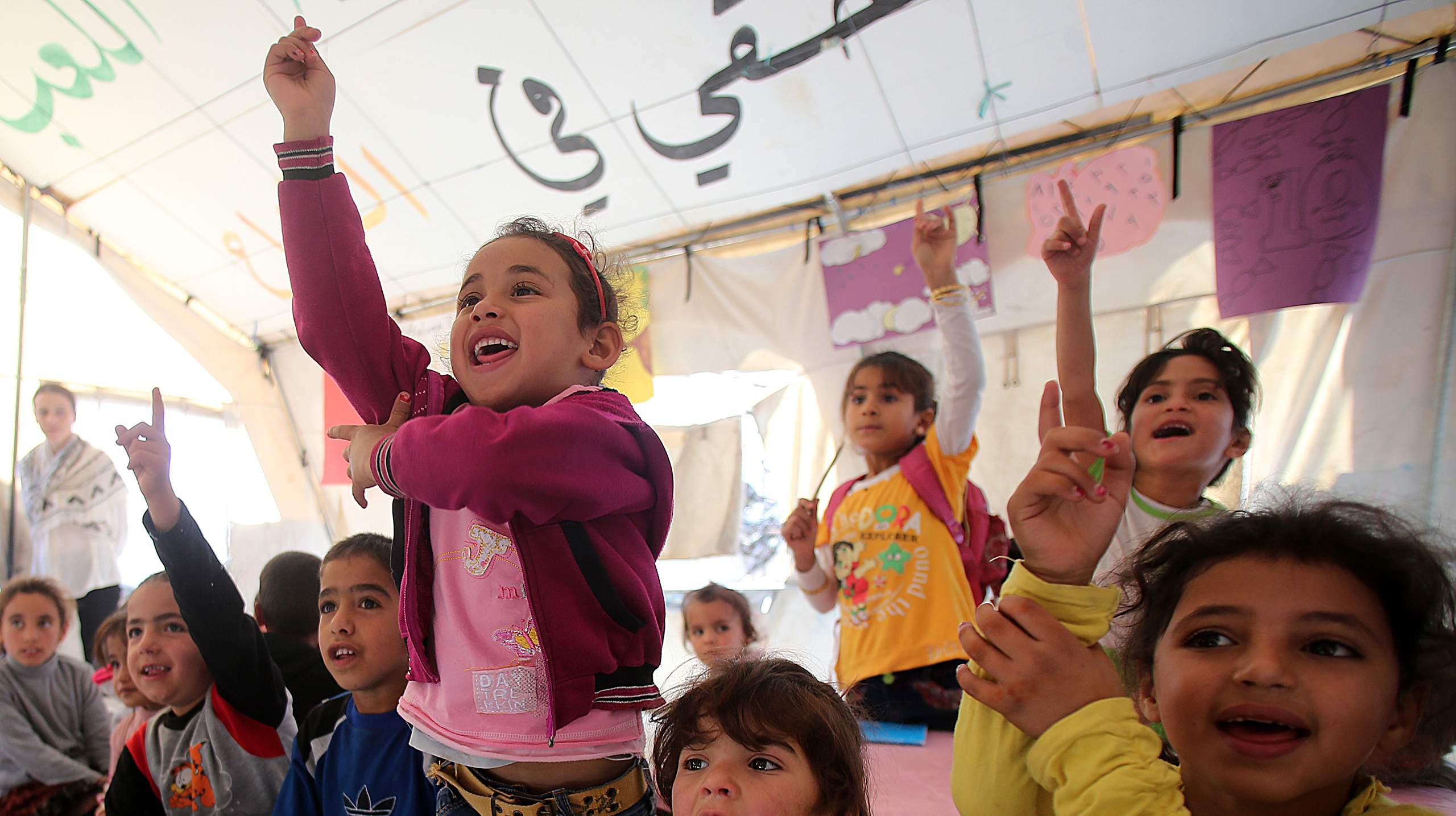 Children in a large tent in a refugee camp. Some have their hands raised.