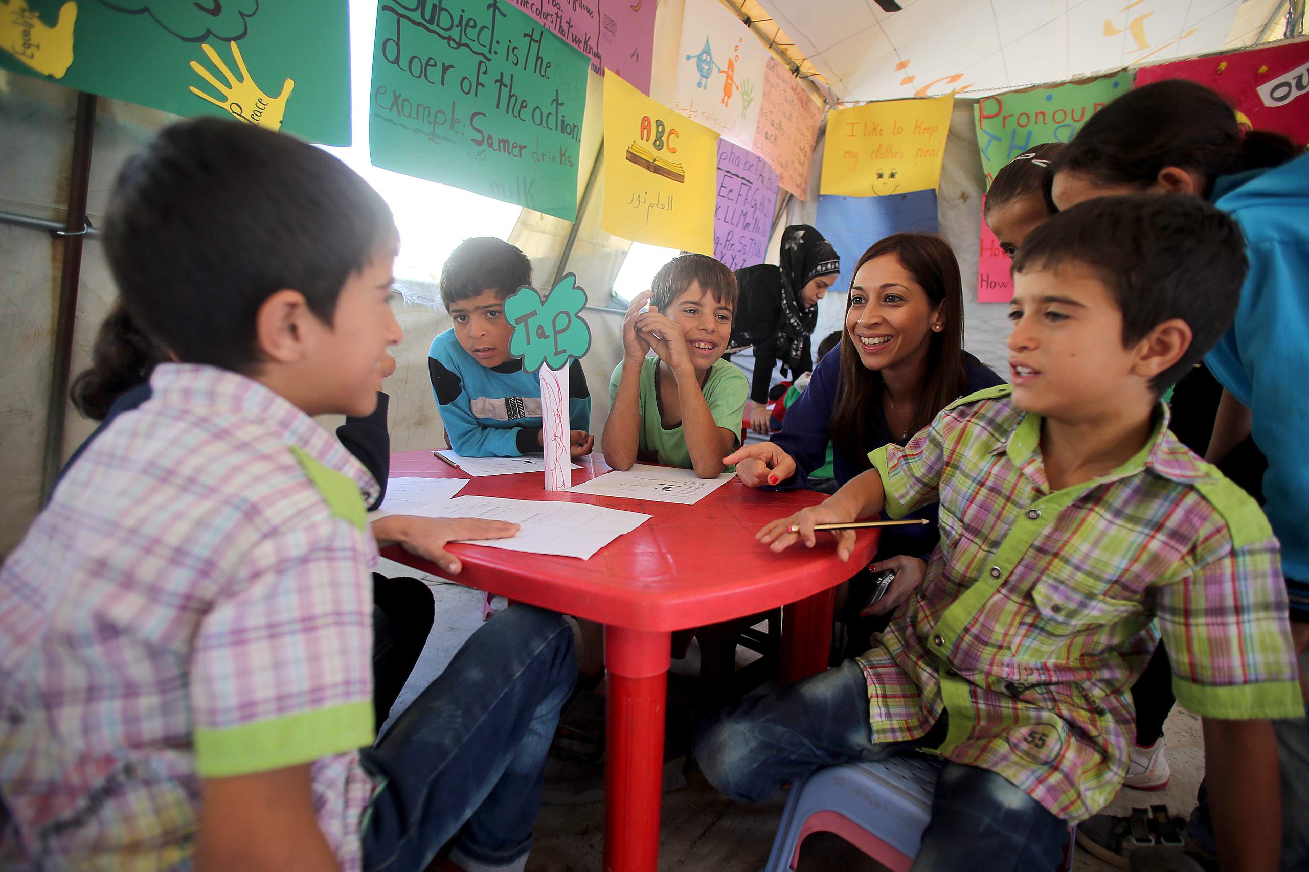 A group of young children talking in a refugee camp. They are sitting at a desk with pencils and paper, and a teacher is looking on.
