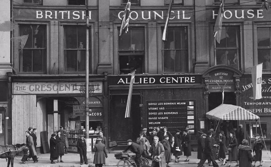 The front of the Allied Centre building. A sign reads welcome in multiple different languages and there are people standing on the street outside. 