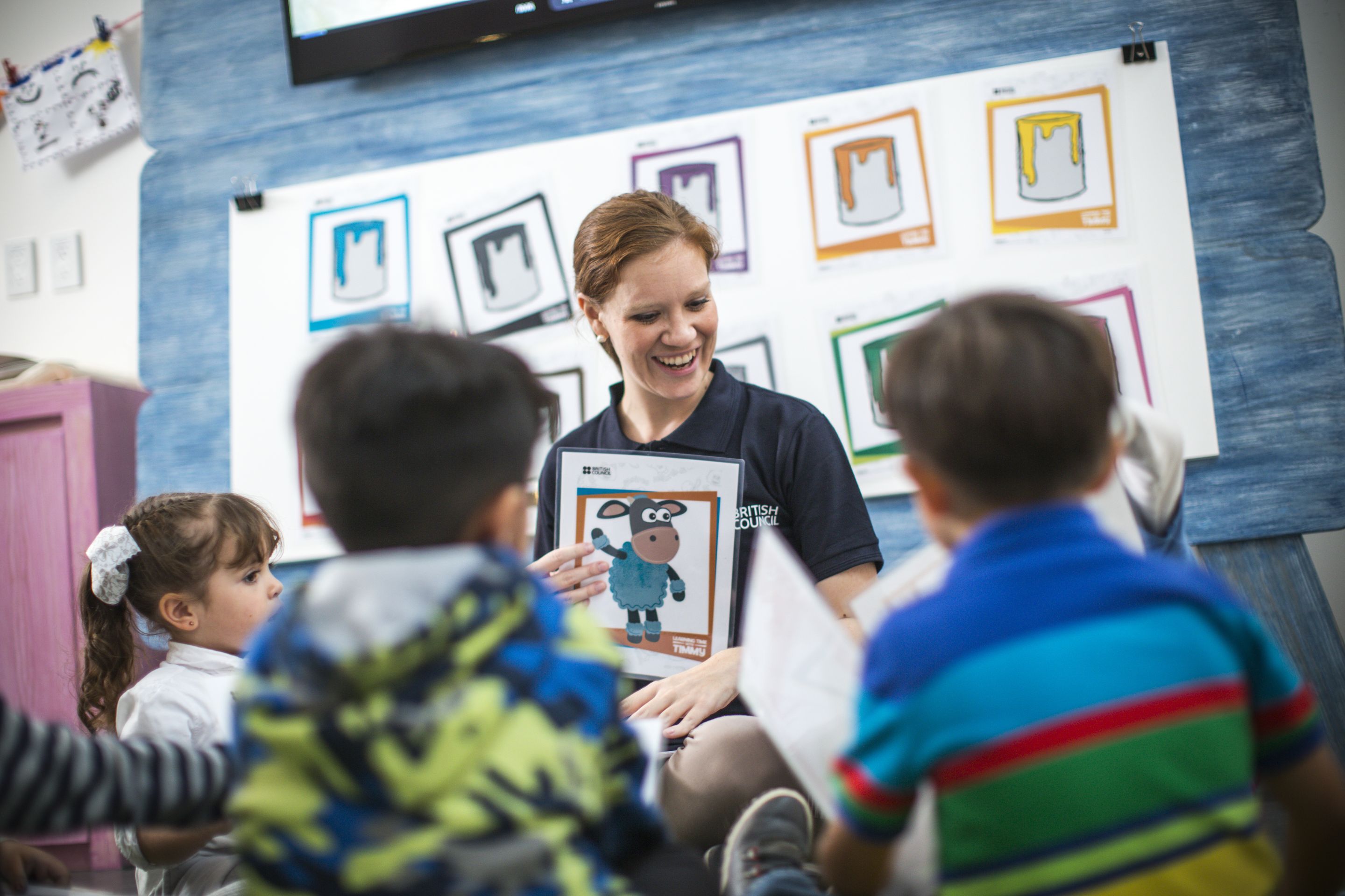 A female teacher holding up a teaching resource with a cartoon sheep on it. She is smiling and a group of young children are sitting with her in a brightly decorated classroom.
