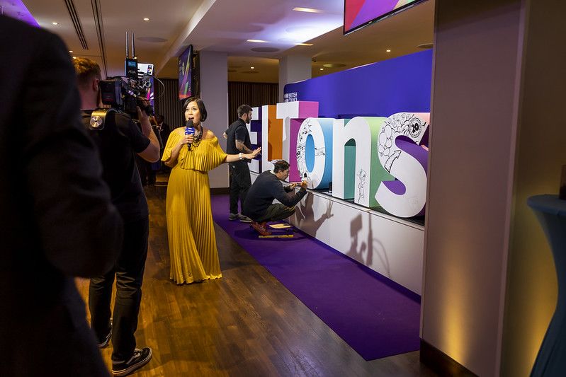 A woman filming a video at ELTons. People are writing on a large ELTons sign behind her.