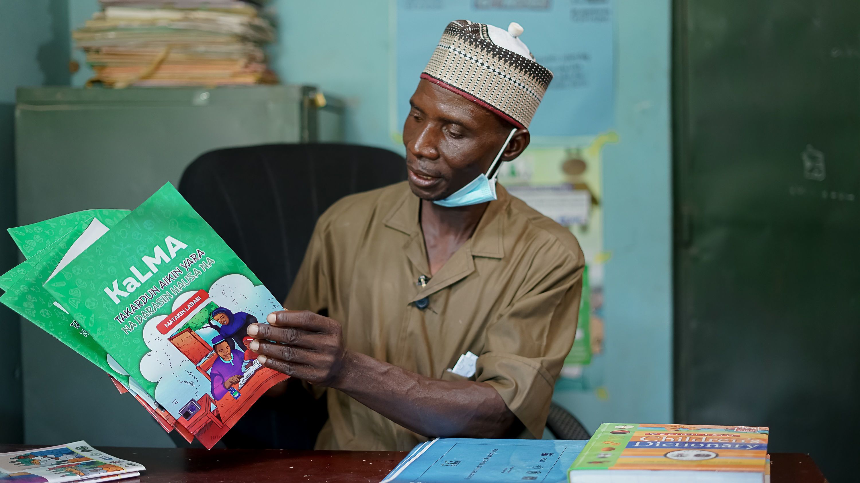 A teacher at a desk looking through a KaLMA workbook written in Hausa.