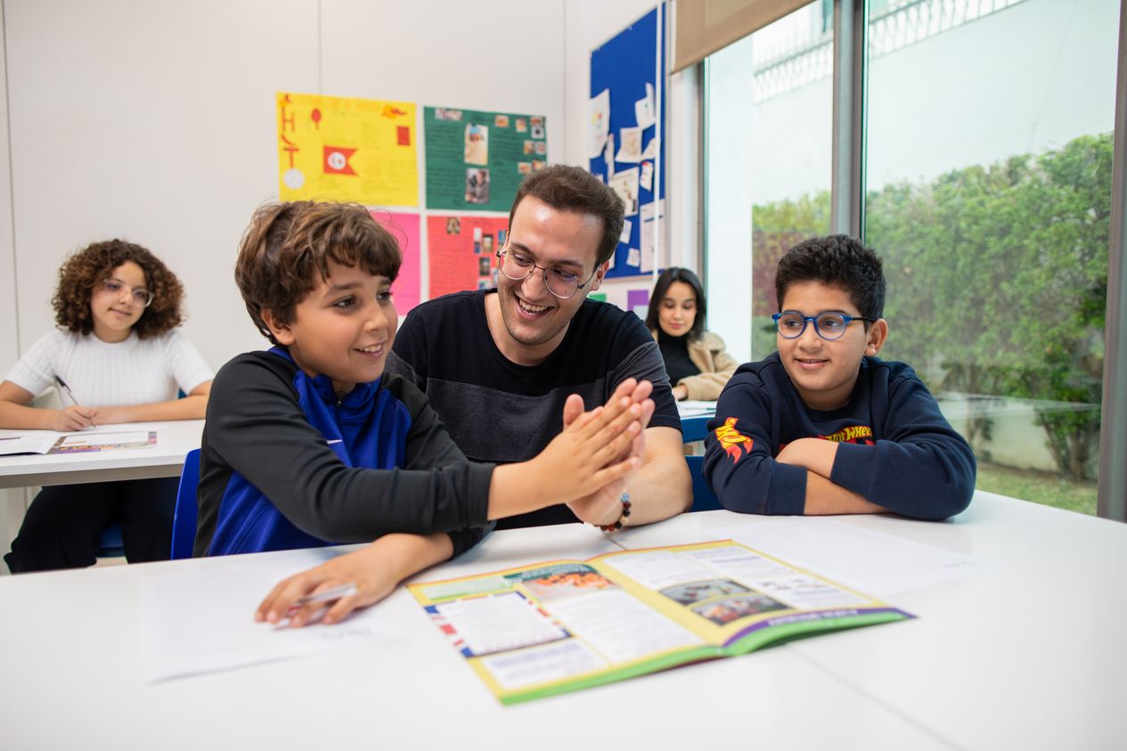 A male teacher high fiving a young boy at a desk in a classroom. They are both smiling.