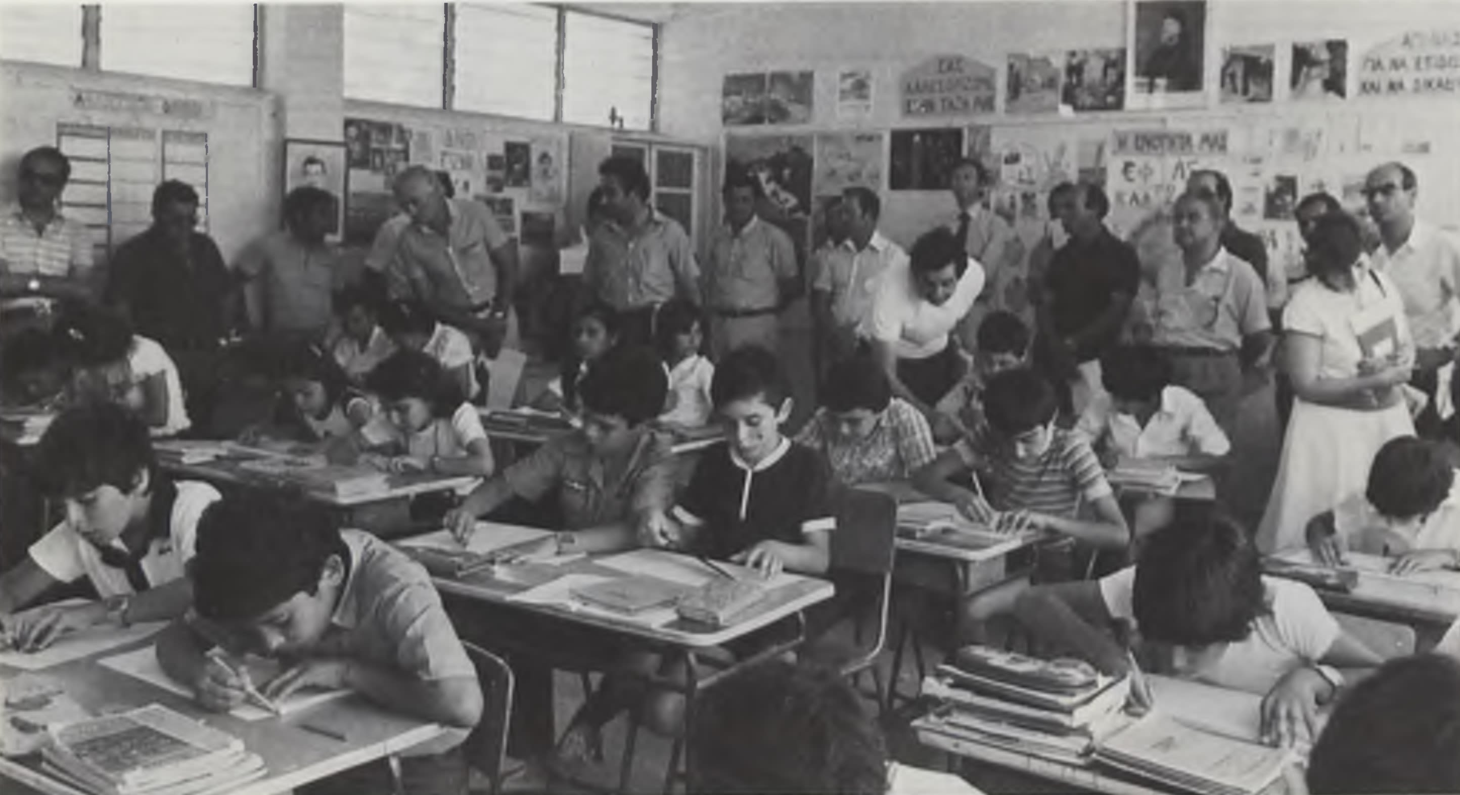 A packed classroom filled by students writing at their desks, observed by a group of teachers standing at the back.