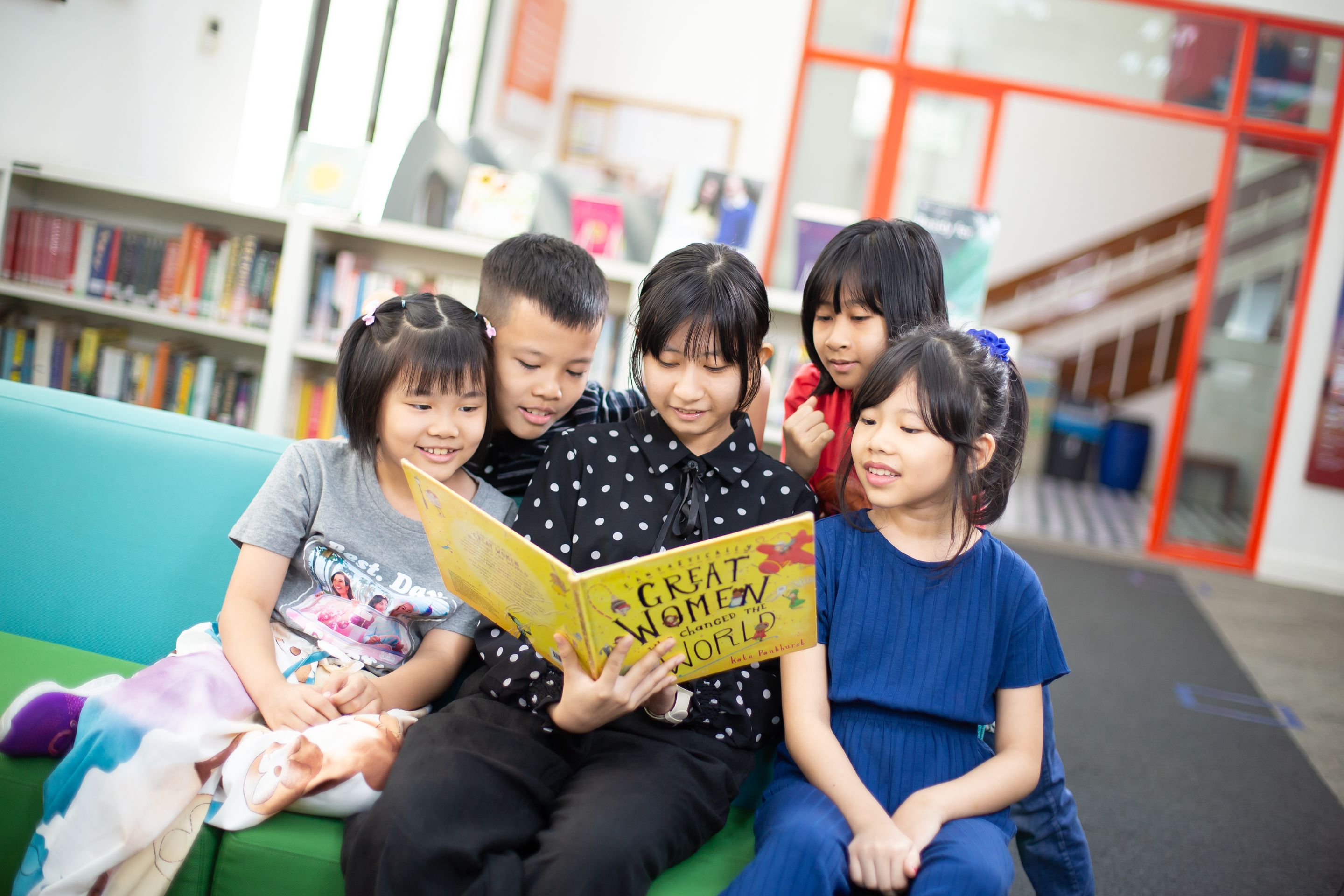 A group of young children reading a book called Great Women who Changed the World in a library. 
