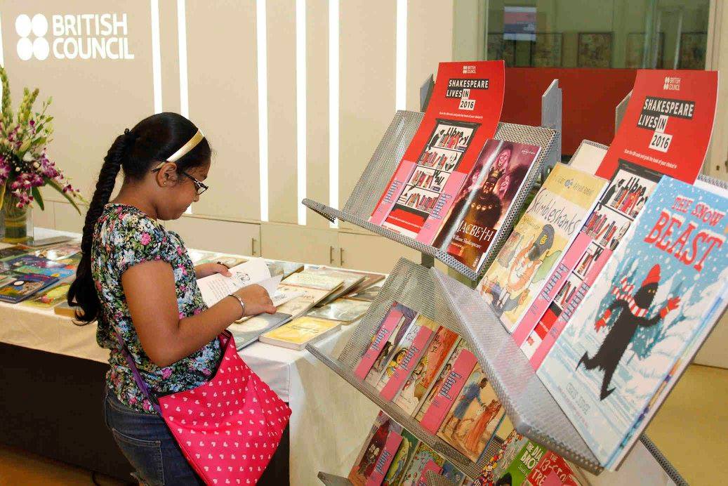 A young girl reading a book at a display with Shakespeare Lives 2016 branding. Children's books and works by Shakespeare are in the display.