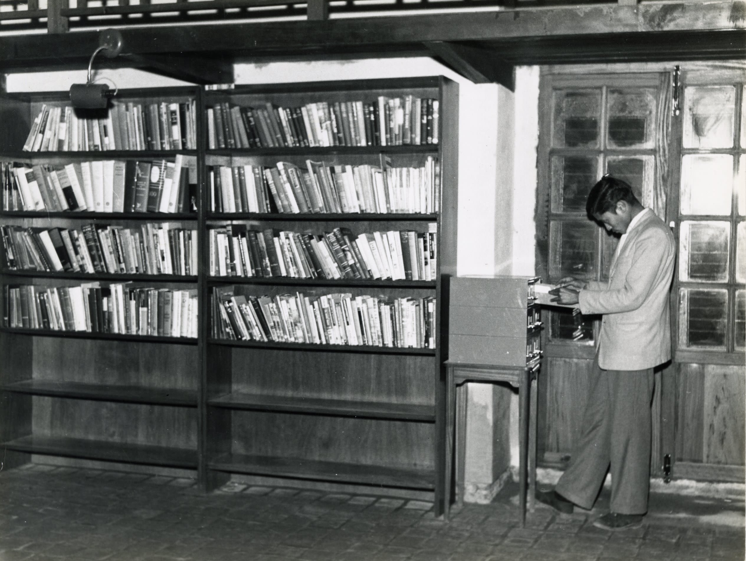 A man looking at something in a drawer. Next to him is a large bookcase with the top four shelves full of books.