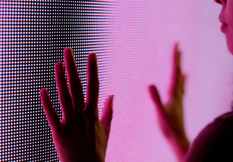 Close-up of a person’s hand touching a vibrant LED screen glowing with pink and purple hues.